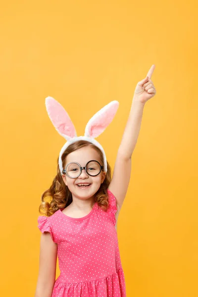 Fröhliches süßes kleines Mädchen in Osterhasenohren und Brille auf gelbem Hintergrund. das Kind zeigt auf eine leere Stelle für Text — Stockfoto