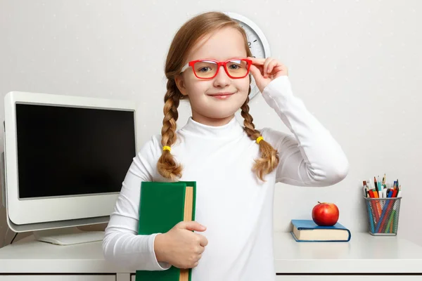 Ragazzina carina con gli occhiali e un libro in mano. Il bambino sta sullo sfondo del posto di lavoro degli studenti — Foto Stock