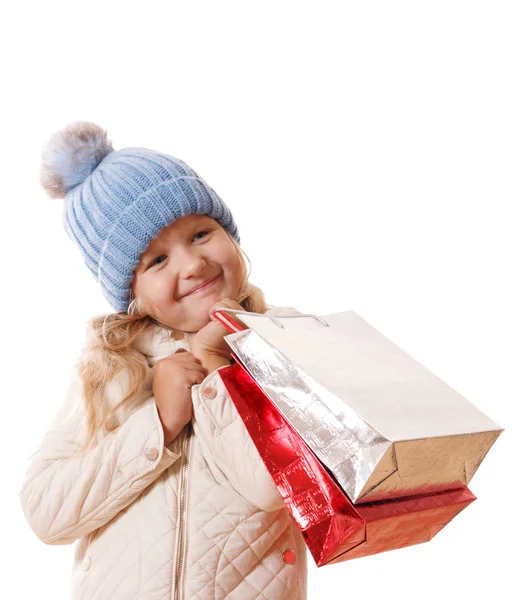 Una niña con un sombrero azul con un pompón y una chaqueta beige con bolsas de papel. El concepto de compras y ventas. Aislado sobre fondo blanco —  Fotos de Stock