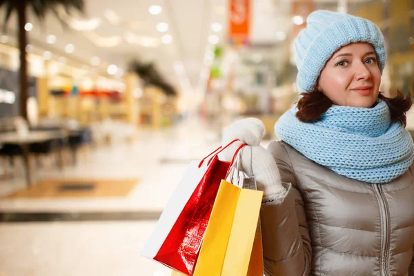 Una donna con un cappello caldo, snood e un piumino, tiene in mano dei sacchetti di carta. Il concetto di shopping e vendite. Al centro commerciale. — Foto Stock