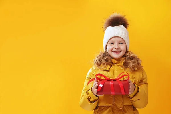 La bambina felice prescolastica in una giacca invernale e in un cappello lavorato a maglia tiene una scatola con un regalo. Il concetto di dare e ricevere doni. Giallo su fondo giallo, rosso — Foto Stock