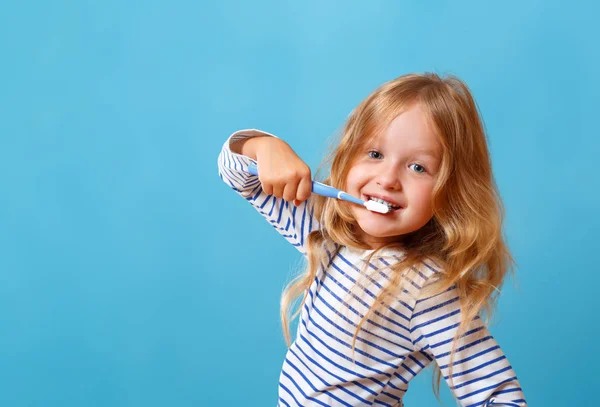 Malá holčička v pruhovaném pyžamu si čistí zuby kartáčkem na zuby. Koncept každodenní hygieny. Izolováno na modrém pozadí — Stock fotografie