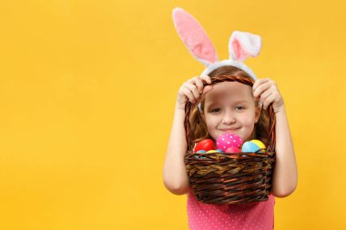Bir tavşanın Paskalya kulaklarında neşeli bir çocuk sepetin sapının altından dışarı bakıyor. Sarı arka planda küçük kızlar