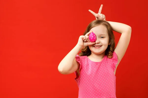 Šťastné dítě s prsty dělá králičí uši a drží růžové velikonoční vajíčko. Portrét malé dívky na červeném pozadí — Stock fotografie