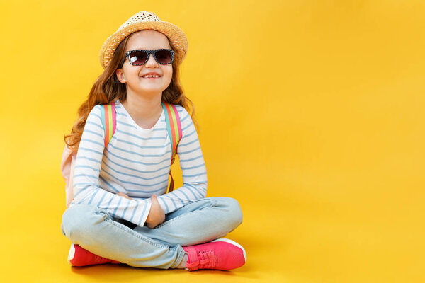 Счастливая маленькая девочка сидит на полу и мечтает о путешествии. Портрет милого красивого ребенка в шляпе, солнцезащитных очках и рюкзаке за спиной на желтом фоне в студии
