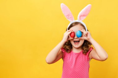 Tavşanın kulaklarında neşeli mutlu çocuk gözlerinde iki Paskalya yumurtası tutuyor. Stüdyoda sarı arka planda küçük bir kızın portresi.