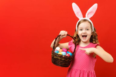 Kırmızı arka planda bir tavşanın kulaklarında tutkulu bir çocuk. Küçük kız paskalya yumurtalarının olduğu sepette parmağını gösteriyor.