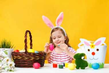 Küçük bir kız bir masaya oturur ve bir fırçayla Paskalya yumurtalarını boyar. Sarı bir duvarın arka planında bir tavşanın kulaklarında mutlu bir çocuk