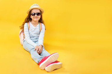 Mutlu moda güzel çocuk stüdyoda yerde oturuyor ve seyahat hayalleri kuruyor. Şapkalı, güneş gözlüklü ve sarı arka planda sırt çantası olan sevimli bir kız. Boşluğu kopyala