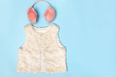 A set of warm clothes. Fluffy pink earmuffs and a white vest on a blue background. Top view flat lay copy space clipart