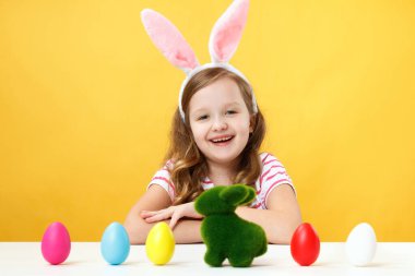 Paskalya konsepti. Tavşan kulaklı mutlu çocuk sarı arka planda. Küçük kız boyalı yumurtalar ve bir tavşanla bir masada oturuyor.