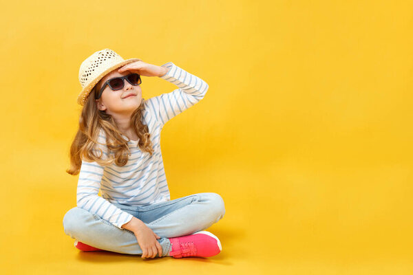 Маленькая девочка сидит на полу в студии в шляпе и солнцезащитных очках на желтом фоне. Ребенок мечтает путешествовать и смотрит вдаль
