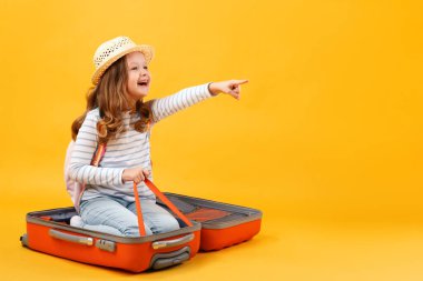 Adventure travel trip dream concept. A cheerful little girl sits inside a suitcase and shows her hand forward. A child on a yellow background in the studio. Copy spac