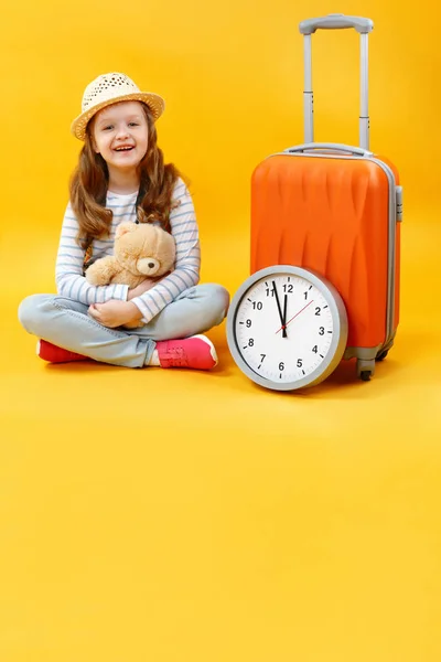 Happy fashionable beautiful baby sits on the floor in the studio and dreams of traveling. Cute little girl with a suitcase, a clock and a soft toy on a yellow background. Copy space
