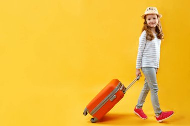 Macera yolculuğu konsepti. Güzel, mutlu küçük kız bir bavulla yürüyor. Stüdyoda sarı arka planda bir çocuğun tam boy portresi. Boşluğu kopyala.