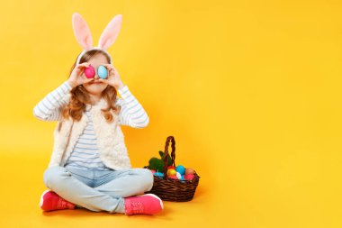 Mutlu Paskalyalar. Neşeli çocuk elinde bir sepet yumurtayla yerde oturuyor. Stüdyoda sarı arka planda tavşan kostümlü şirin bir kız. Boşluğu kopyala.