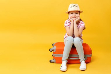 Küçük kız bir bavula oturur ve sarı arka planda avuçlarıyla yanaklarını tutar. Bir çocuk yolculuk, yolculuk, macera, rahatlama için bekliyor..