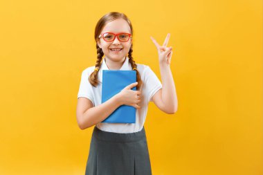 Sarı arka planda gözlüklü küçük bir kız çocuğunun portresi. Çocuk elinde bir kitap tutuyor ve bir zafer işareti gösteriyor. Eğitim, başarı ve okul konsepti. Boşluğu kopyala.