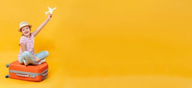 Küçük mutlu bir kız bavulda oturuyor ve sarı arka planda kağıttan bir uçak tutuyor. Çocuk seyahat ve rahatlama hayali kuruyor. Boşluğu kopyala.