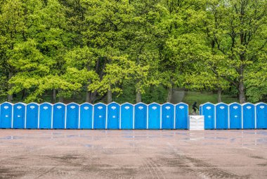 Long row of portable toilets in front of a forest where one has tipped over. clipart