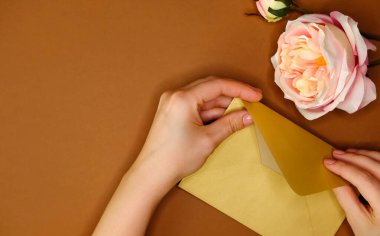 Altın zarf, yapay pembe gül ve altın konfeti simli kahverengi arka planda kadın eli. Düzleştirme biçimi. 