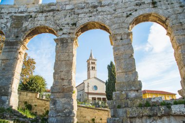 Hırvatistan 'ın Pula kentindeki antik miras. Katolik kilise kulesi çanı, anıtsal antik Roma arenasının kemerlerinden görünüyor. Tarihi amfi tiyatronun içi, mavi arka plandaki yüksek duvarların geniş açılı görüntüsü..