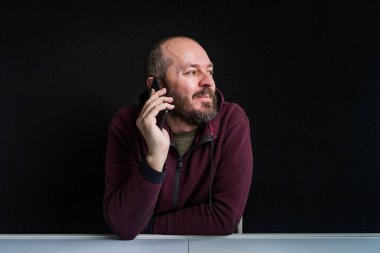 Beyaz masanın arkasında oturan beyaz sakallı, gülümseyen ve siyah arka planda izole akıllı telefondan konuşan beyaz adamın stüdyo fotoğrafı.