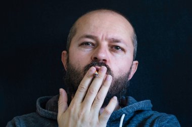 Kapüşonlu, sakallı, sigara içen, sağlıksız bir alışkanlık, nikotin bağımlısı kafa ve omuz resmi olan yetişkin bir adam.
