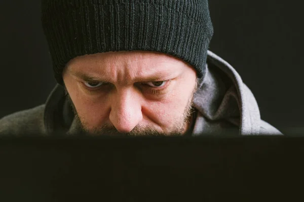 Man wearing black cap and hoodie sitting behind the computer monitor in the dark, looking at computer screen typing, close up, hacking computer system concept