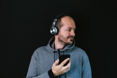 Sakallı, kapüşonlu, siyah arka planda duran, kulaklıklı, müzik dinleyen ve gülümseyen kafa ve omuzlu rahat bir adam.