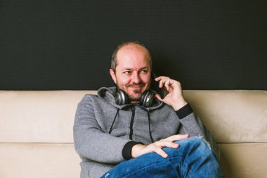 Sakallı, rahat, kapüşonlu ve pantolonlu bir adam kanepeye oturmuş, kulaklıkları boynunda ve akıllı bir telefonla konuşuyor.
