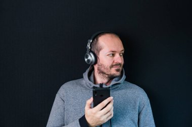 Sakallı, kapüşonlu, siyah arka planda duran, kulaklıklı, müzik dinleyen ve gülümseyen kafa ve omuzlu rahat bir adam.