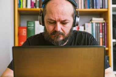 Ev işi konsepti. Yetişkin sakallı adam internetten bilgisayarlı bilgisayarda çalışıyor, arkasında kitap rafları var.