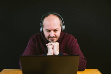 Kapüşonlu ve kulaklı bir adam dizüstü bilgisayarın arkasında oturuyor, kafası karışmış, başarısız, siyah arka planda.