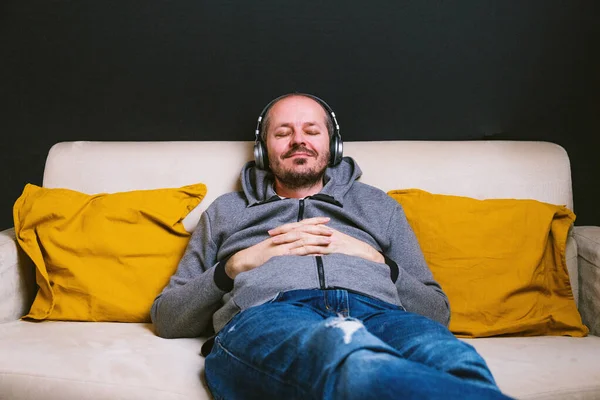 Bearded man in hoodie and trousers relaxed couch with headphones and listening music, smiling, eyes closed, resting at home