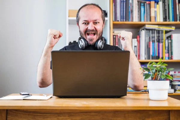 Man working from home office, happy and cheerful. Good news, new job. Bearded man on computer laptop behind vintage desk with flower in vase, with headphones, celebrating good news with colleagues on online meeting.