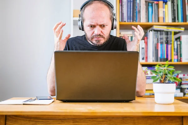 Man working from home office, mad at someone or something. Bearded man with headphones sitting behind vintage desk at home, angry face, having business problem