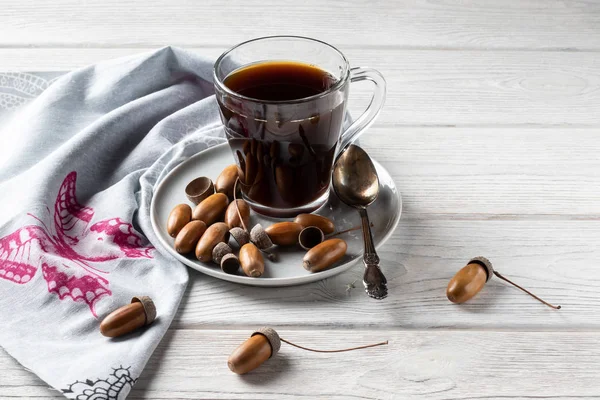Hot coffee made from acorns in a glass with a napkin is a tonic drink with a coffee flavor, rich color and pleasant aroma. On a white wooden background — Stock Photo, Image