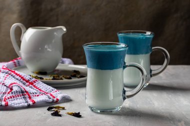 2 bardak mavi Dalgona Matcha Blue Latte, açık gri arka planda bezelye kelebeklerinden krema içeceği. Yatay yönelim