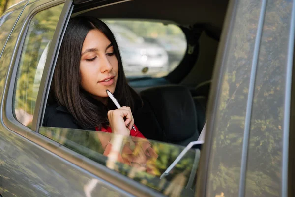 Stylish woman touching pen to her chin in car