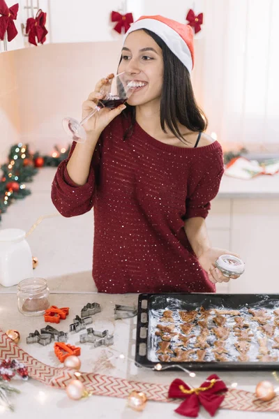 Pretty woman with Santa hat drinking wine and making Christmas cookies.