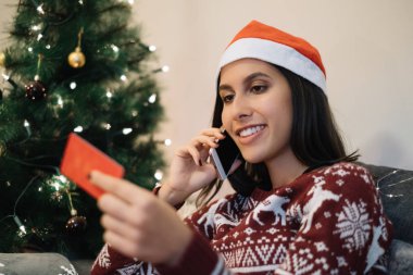 Gülümseyen kız Noel ağacının yanında telefonla konuşuyor.