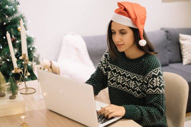 Noel Baba şapkalı güzel bir kadın dizüstü bilgisayarın önünde oturuyor.