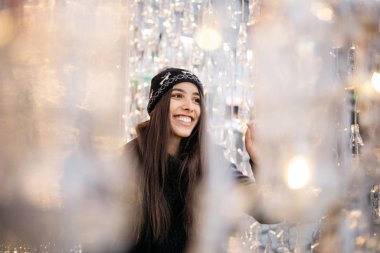 Perde ışığı ve şişelerle çevrili esmer kız gülümsüyor.