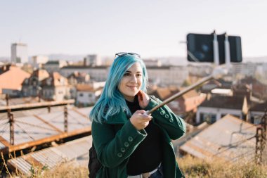 Mavi saçlı, gülümseyen ve şehirde selfie çeken bir kız.