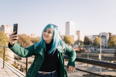 Kadın şehir manzaralı çitlere yaslanırken selfie çekiyor.