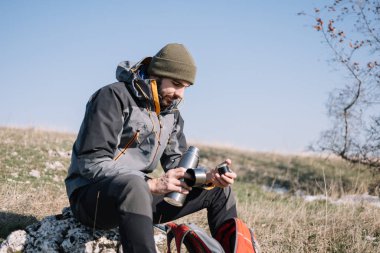 Erkek yürüyüşçü bir kayanın üzerinde oturuyor ve termosu açıyor.