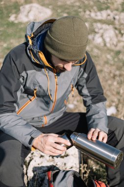 Bir kayanın üzerinde oturan yürüyüşçü termostan kahve dolduruyor.