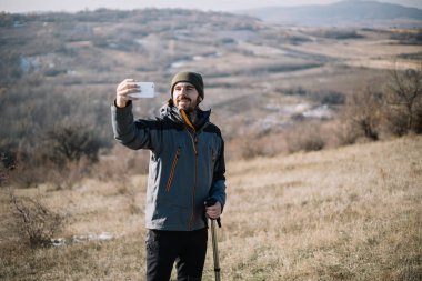 Sakallı yürüyüşçü, dağ manzaralı akıllı telefon kullanarak selfie çekiyor.