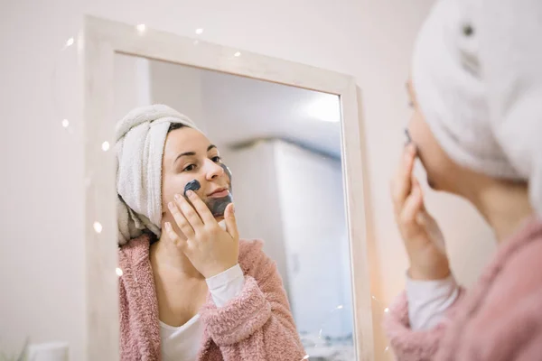 Chica joven aplicando máscara en su cara delante de un espejo — Foto de Stock